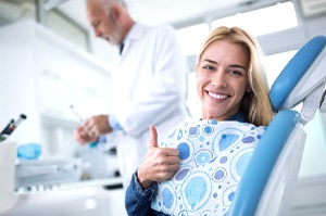 A young woman sitting upright in the dentist’s chair and giving a thumbs up while the dentist prepares to perform air abrasion