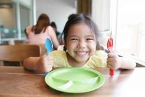 child smiling and sitting at a table with a fork and spoon in their hands