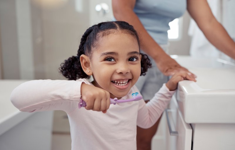 Young girl smiling with teeth holding her mother's hand in one hand and a toothbrush with toothpaste on it in the other in front of a bathroom sink